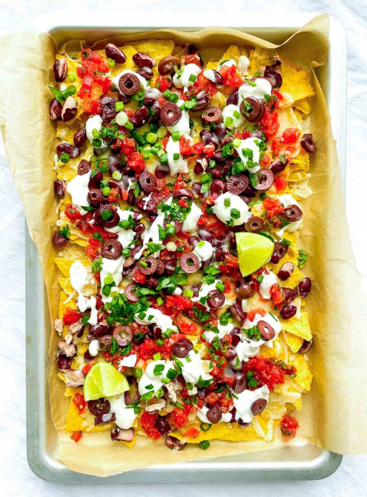 Sheet pan nachos loaded with melted cheese, kidney beans, salsa, sliced olives, sour cream, chopped green onions, and lime wedges.