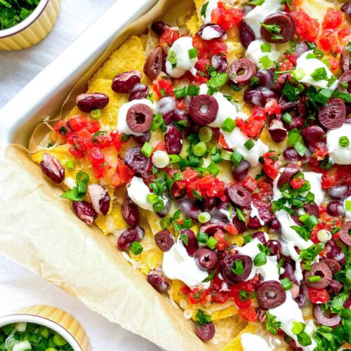 Close-up of loaded sheet pan nachos with melted cheese, kidney beans, salsa, sliced olives, sour cream, and chopped green onions.