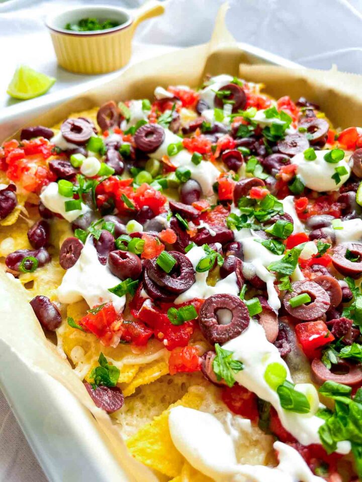 Side view of loaded sheet pan nachos with melted cheese, kidney beans, salsa, sliced olives, sour cream, and chopped green onions.