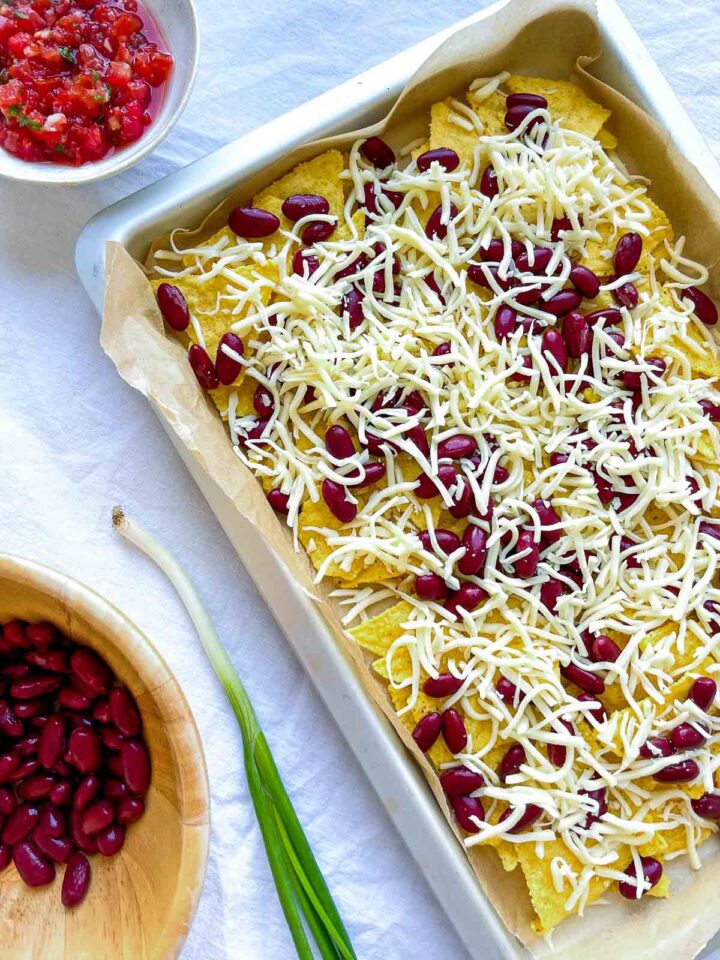 Sheet pan nachos with tortilla chips topped with shredded cheese and red kidney beans, ready to bake, with salsa and green onions nearby.