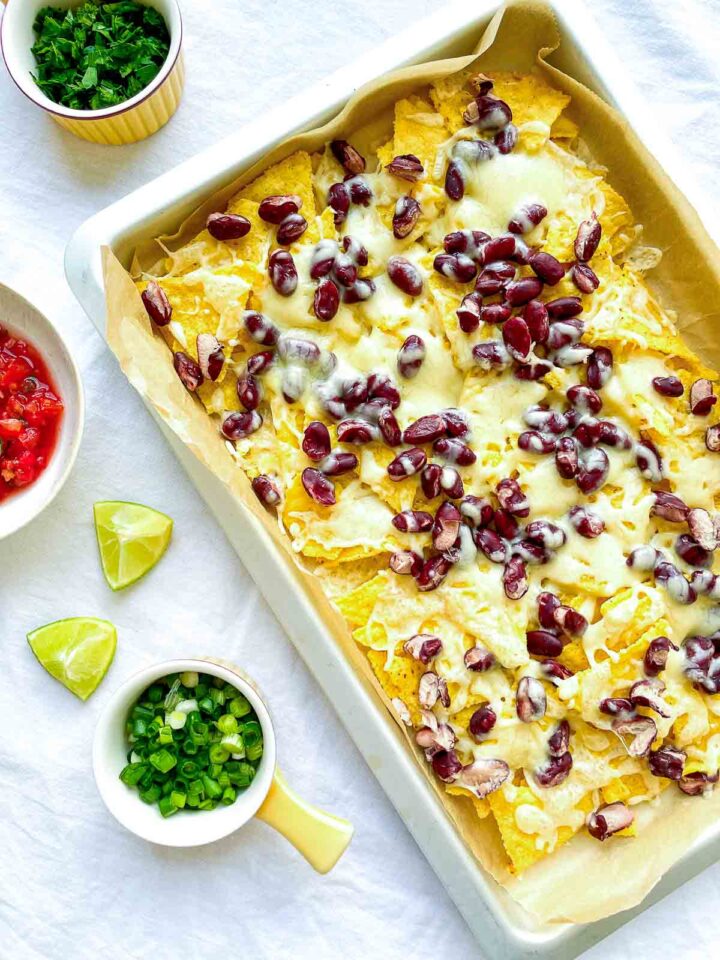 Sheet pan nachos topped with melted cheese and red kidney beans, served with salsa, lime wedges, and chopped green onions.