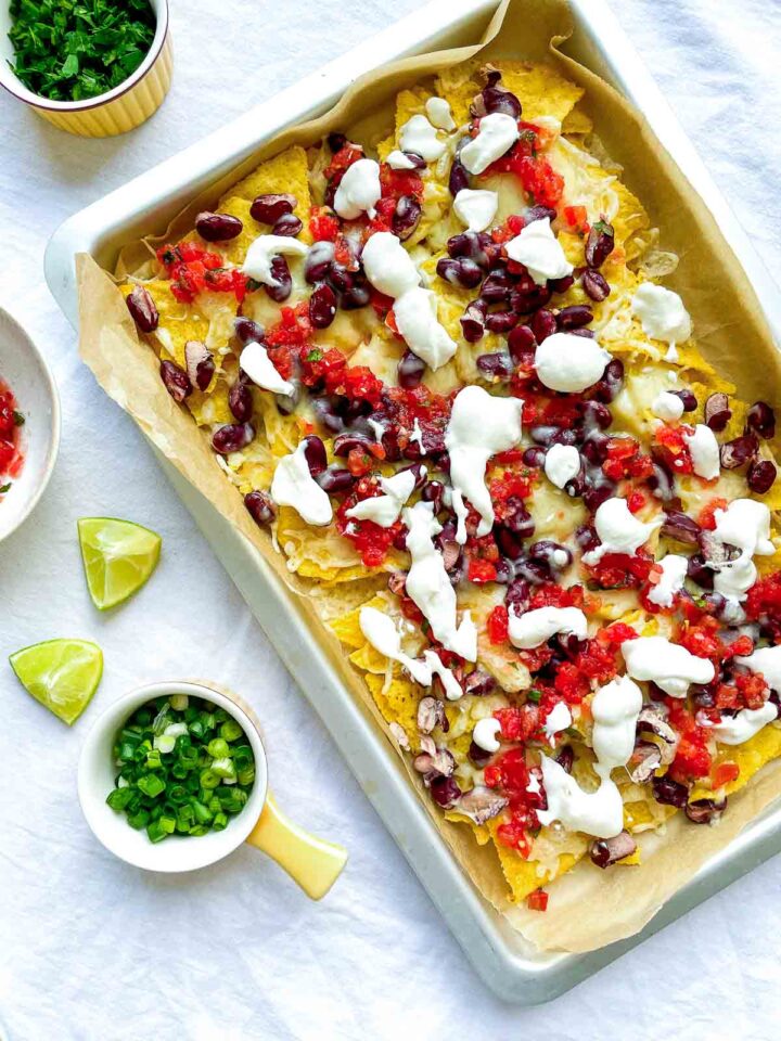 Sheet pan nachos topped with melted cheese, kidney beans, salsa, and sour cream, served with lime wedges and chopped green onions on the side.