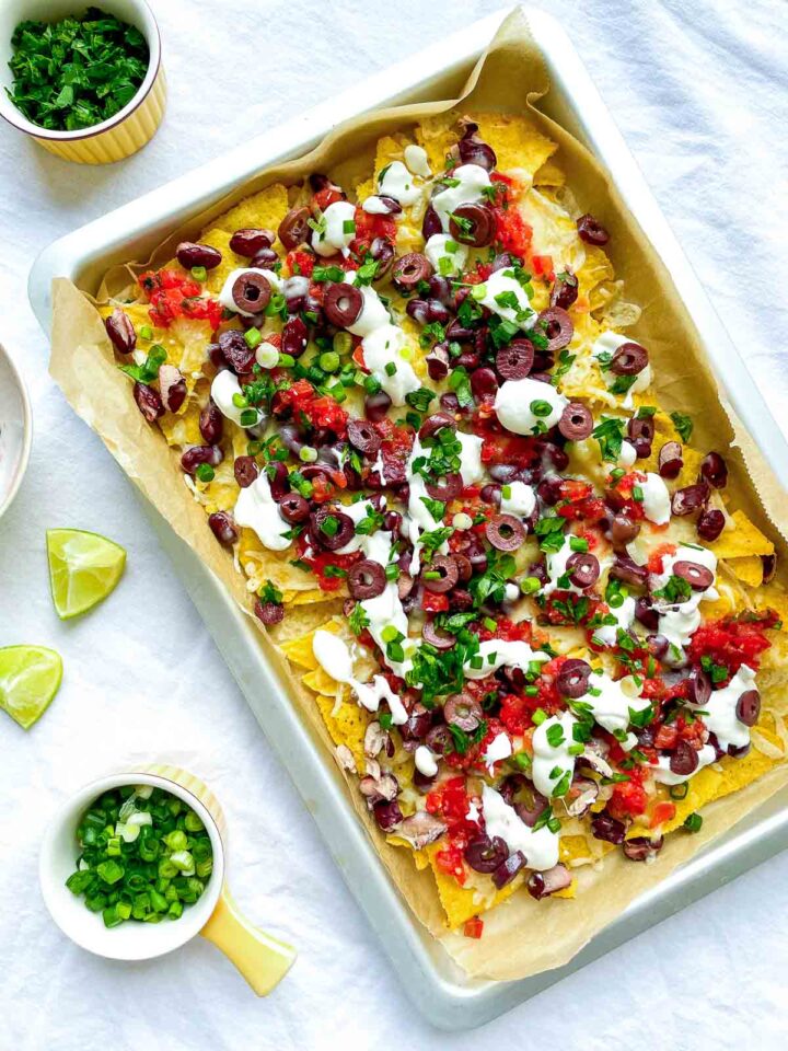 Sheet pan nachos topped with melted cheese, kidney beans, salsa, sliced olives, sour cream, and chopped green onions, served with lime wedges.