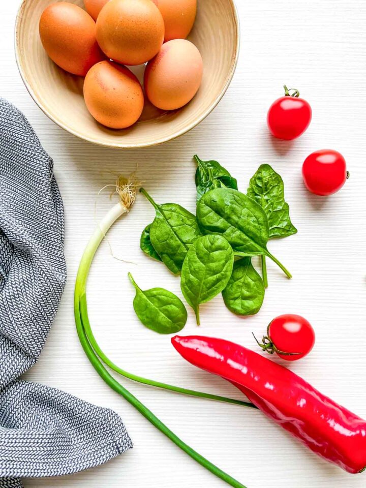 Bowl of eggs with fresh spinach, cherry tomatoes, green onion, and red pepper on a countertop.