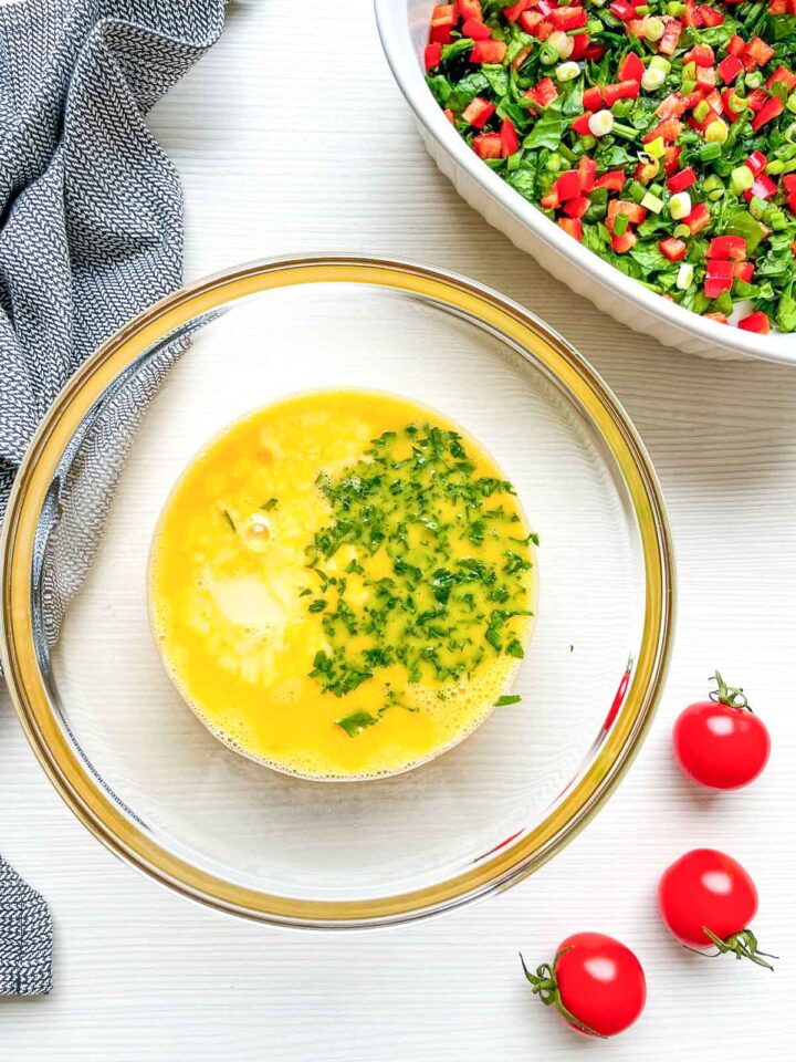 Bowl with beaten eggs and herbs next to chopped spinach and vegetables.