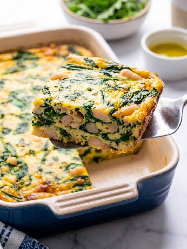 A square baking dish filled with a spinach and white bean egg bake, with a thick square slice lifted on a serving spatula showing whole white beans layered throughout the fluffy egg and greens, set on a marble countertop with a small bowl of olive oil and fresh greens in the background.