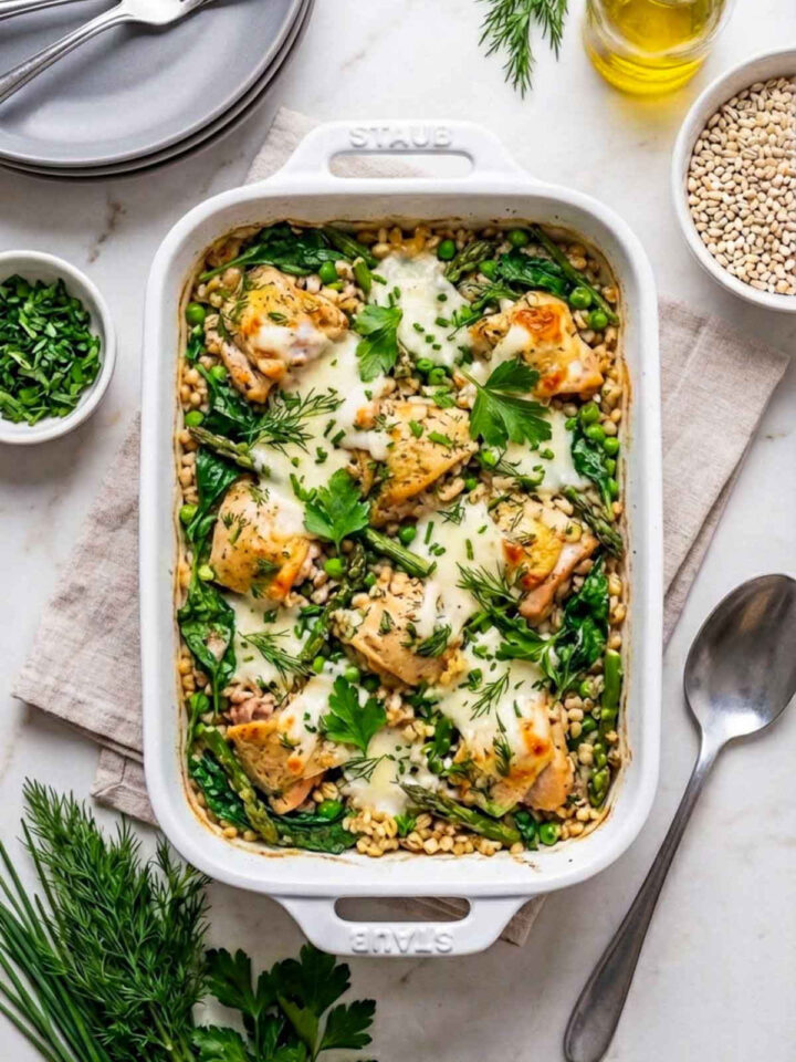 A white ceramic baking dish filled with a spring herb chicken and barley casserole topped with melted cheese, tender chicken pieces, asparagus, peas, and fresh herbs, surrounded by bowls of barley, olive oil, dill, parsley, and plates on a marble countertop.