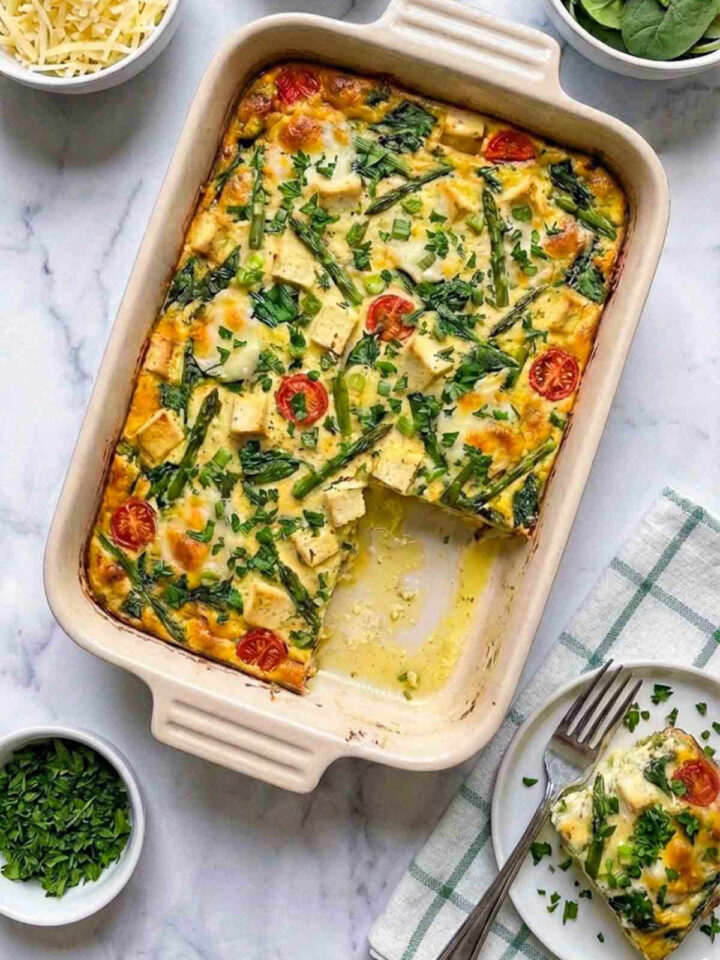 A baked spring vegetable and tofu breakfast casserole with asparagus, spinach, cherry tomatoes, and tofu cubes in a ceramic dish with a slice served on a plate.