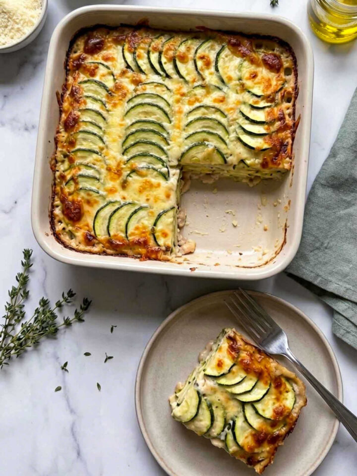 A square ceramic baking dish filled with layered zucchini rounds baked in a creamy white bean sauce and topped with golden melted cheese, with one square removed and plated below showing the tender zucchini layers and creamy interior, surrounded by bowls of white beans, grated cheese, olive oil, and fresh thyme on a marble surface.