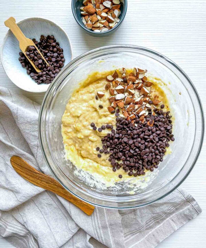 Banana bread batter mixed with flour, topped with chocolate chips and chopped almonds, ready to be folded together for a rich, nutty bake.