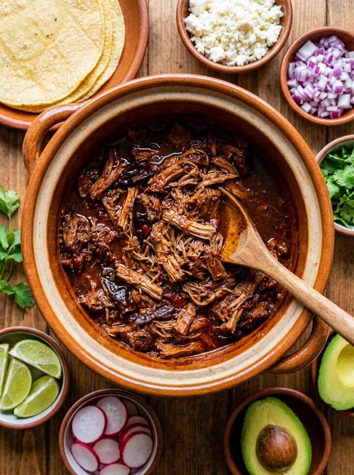 Ancho chile shredded pork in a crockpot with a rich dark sauce, served with tortillas, onion, cilantro, lime, and avocado on a wooden surface.