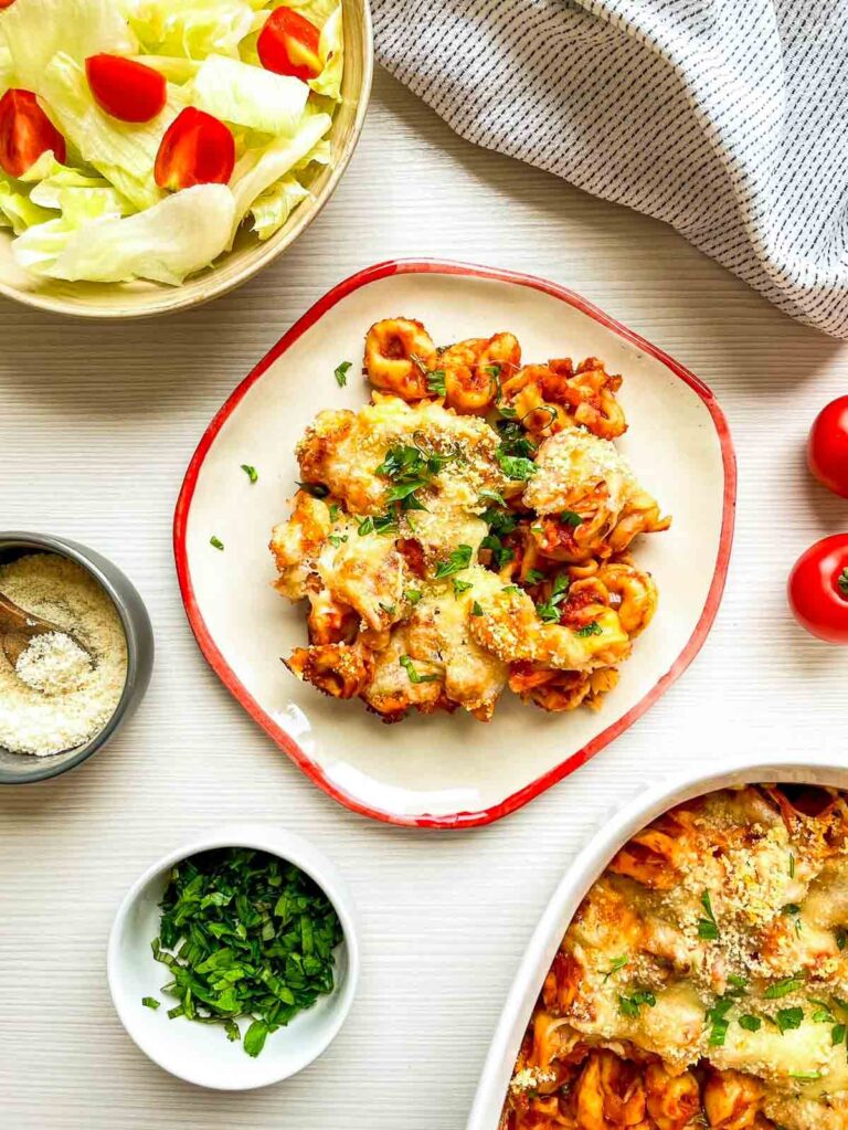 Cheesy baked tortellini topped with herbs and parmesan, served with a fresh side salad and cherry tomatoes on a light table setting.