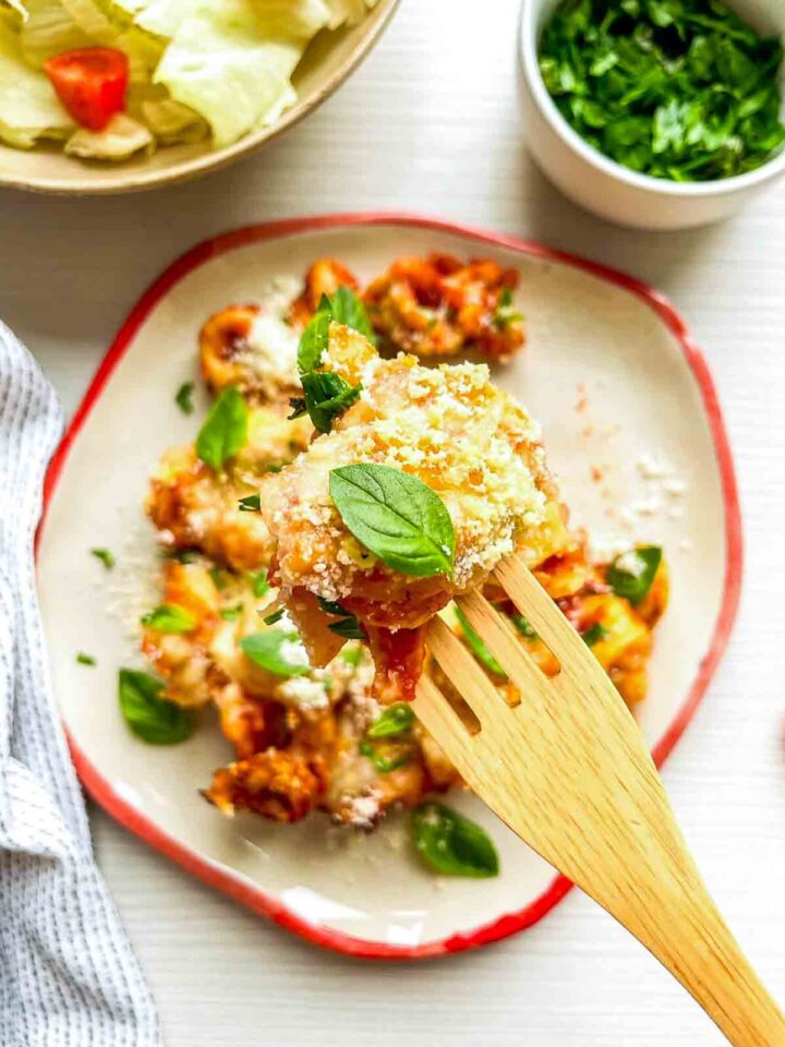A forkful of cheesy baked tortellini topped with parmesan and fresh basil, showing the rich, saucy layers up close