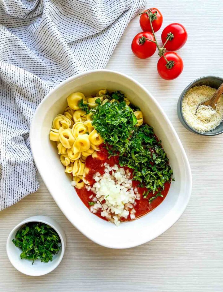 Tortellini layered with marinara sauce, chopped onions, and fresh herbs, ready to be mixed and baked.