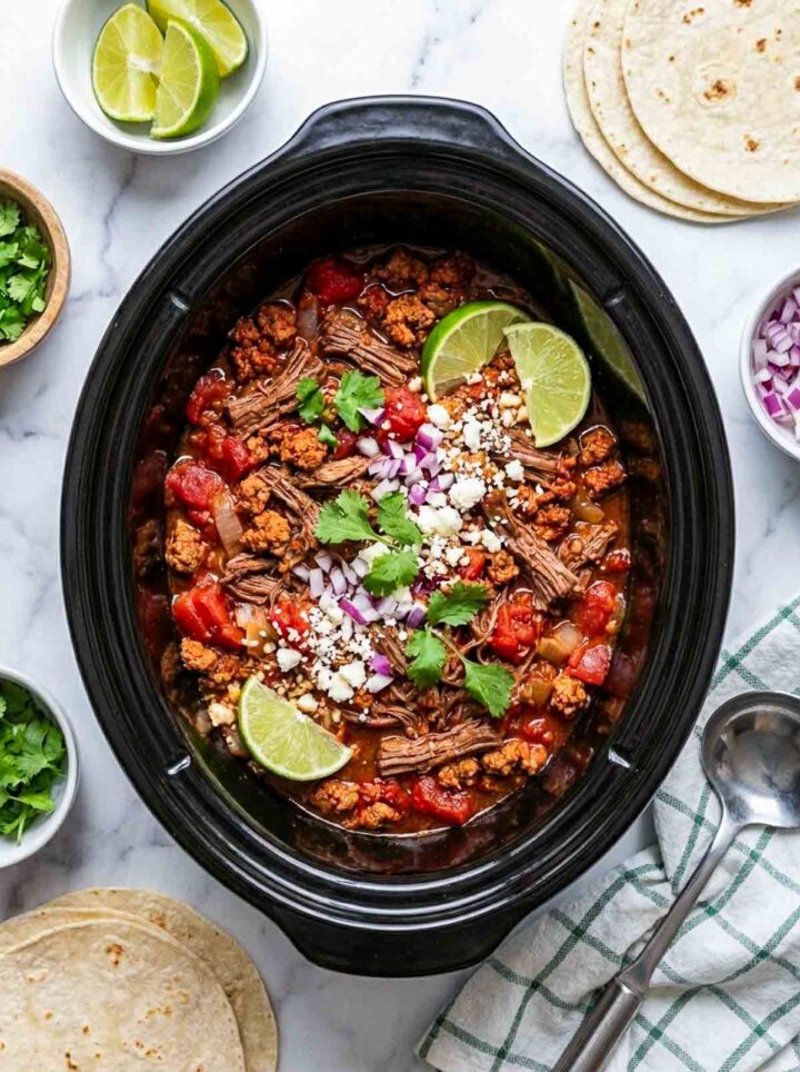 Crockpot beef and chorizo taco filling with tomatoes, topped with cilantro, onion, and cotija cheese, served with tortillas and lime on a marble surface.