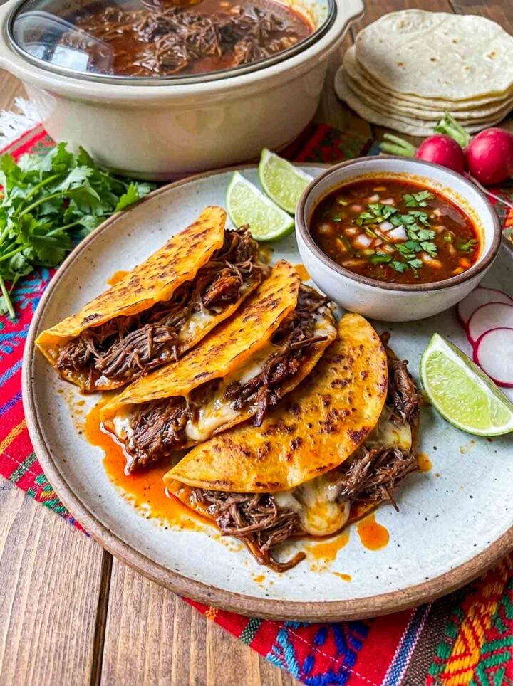 Crispy birria beef tacos filled with shredded meat, served with dipping broth, lime wedges, and cilantro on a plate with a slow cooker in the background.