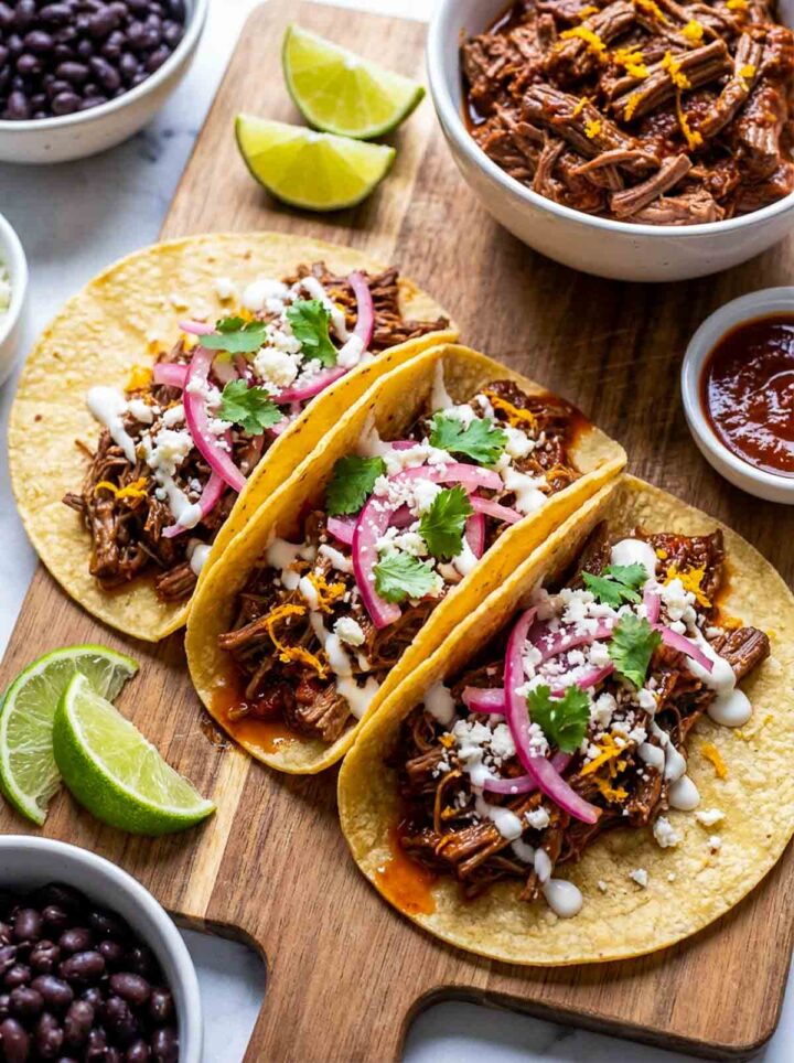 Chipotle orange shredded beef tacos topped with pickled onions, cotija cheese, cilantro, and crema, served with lime wedges and beans on a wooden board.