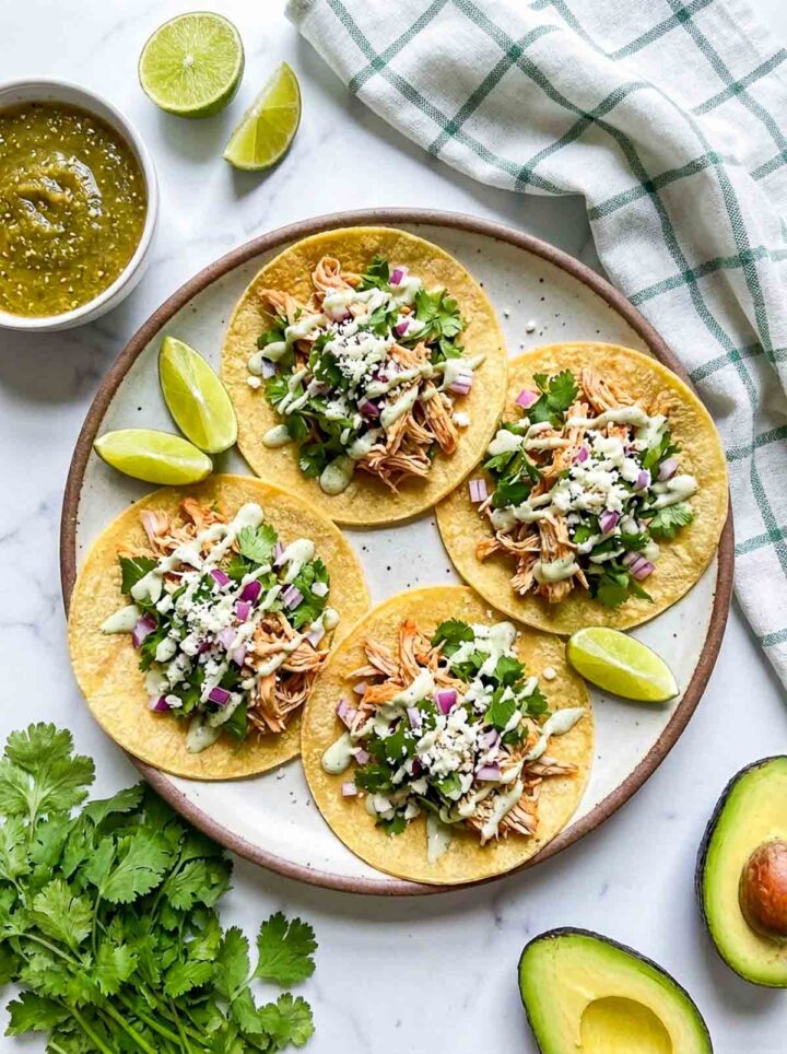 Cilantro lime shredded chicken street tacos topped with cotija cheese, onion, and creamy sauce, served with lime wedges, avocado, and salsa on a marble surface.