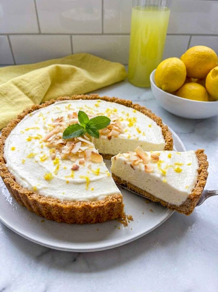 A frozen limoncello coconut cream pie with a biscotti crust, topped with toasted coconut, lemon zest, and mint, with one slice being lifted from the pie on a white plate, lemons and a glass of lemonade in the background.