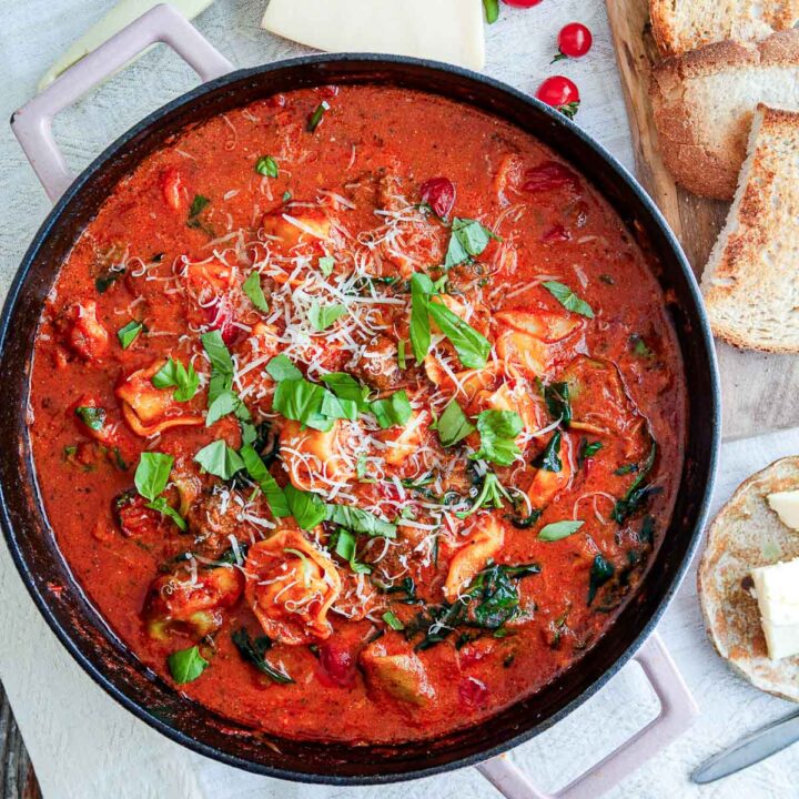 Finished Italian sausage tomato skillet tortellini in a large dark pan garnished with fresh basil leaves and grated parmesan, served alongside crusty bread.