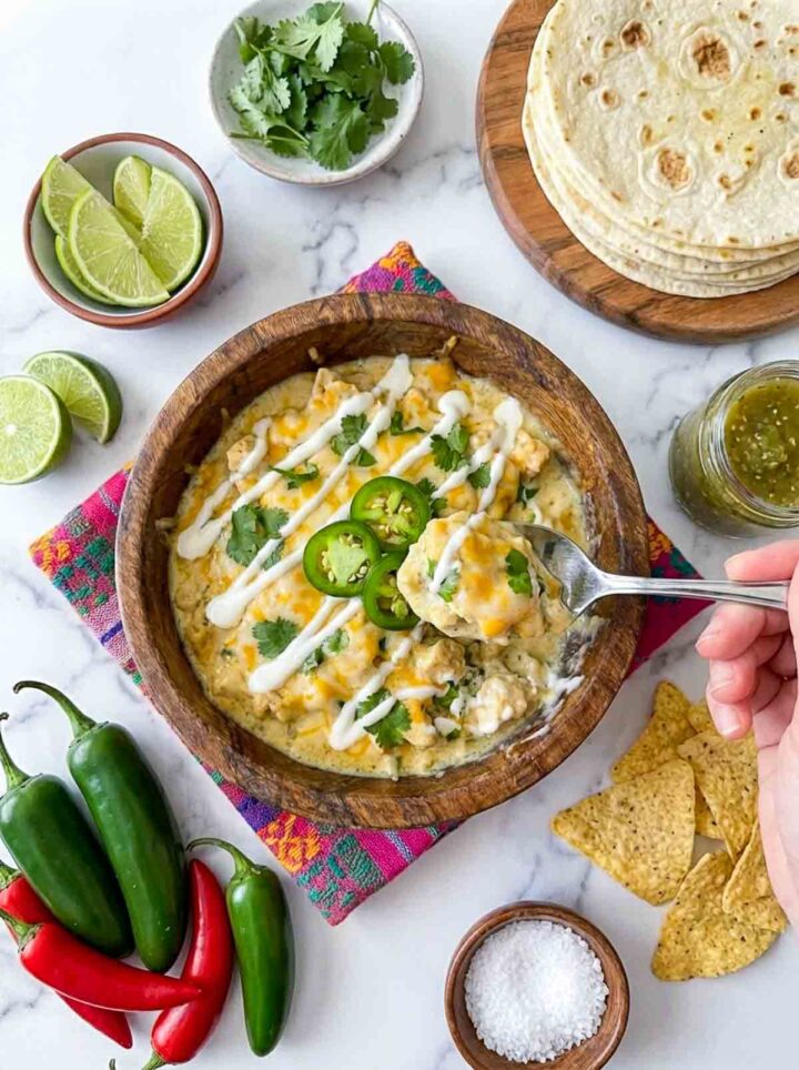 Creamy jalapeno chicken enchilada filling in a bowl topped with cheese, sliced jalapenos, and crema, served with tortillas and chips on a marble surface.