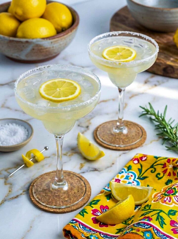 Two limoncello lemonade margaritas in stemmed glasses with salted sugar rims and lemon slices, served on a marble surface with a patterned yellow napkin and fresh lemon wedges.