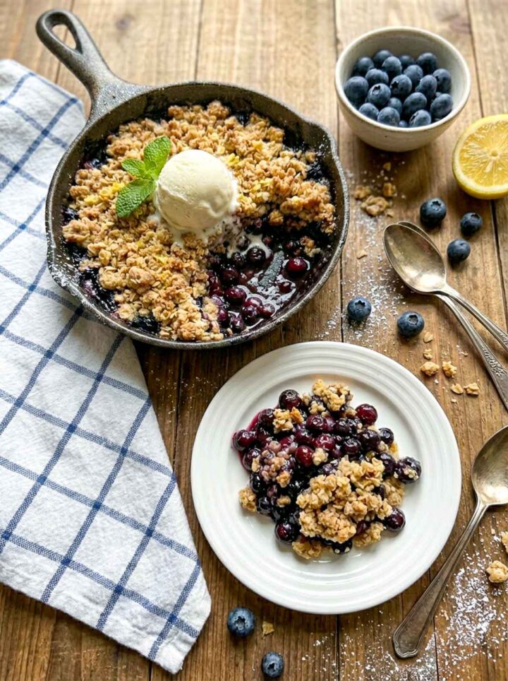 A blueberry crumble baked in a cast iron skillet topped with a scoop of vanilla ice cream, served with a plated portion, fresh blueberries, lemon, and a blue check napkin on a rustic wooden surface.
