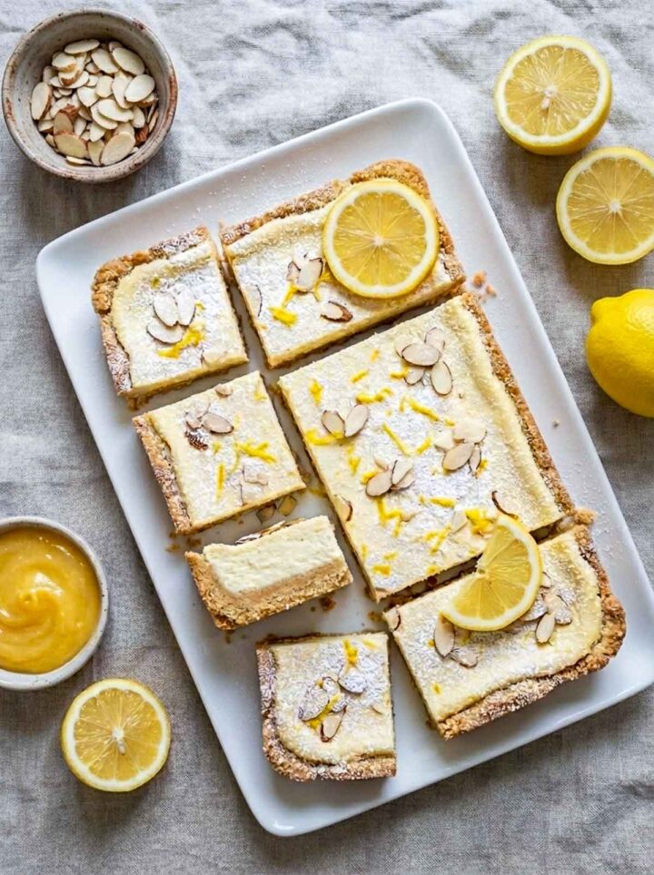 Limoncello cheesecake bars with almond shortbread crust dusted with powdered sugar and topped with sliced almonds and lemon zest, served on a white plate with lemon halves and a yellow napkin on a marble surface.