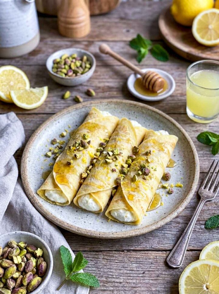 Rolled limoncello mascarpone crepes filled with creamy mascarpone, drizzled with honey and topped with chopped pistachios, served on a rustic plate with lemons, mint, and a honey dipper on a wooden surface.