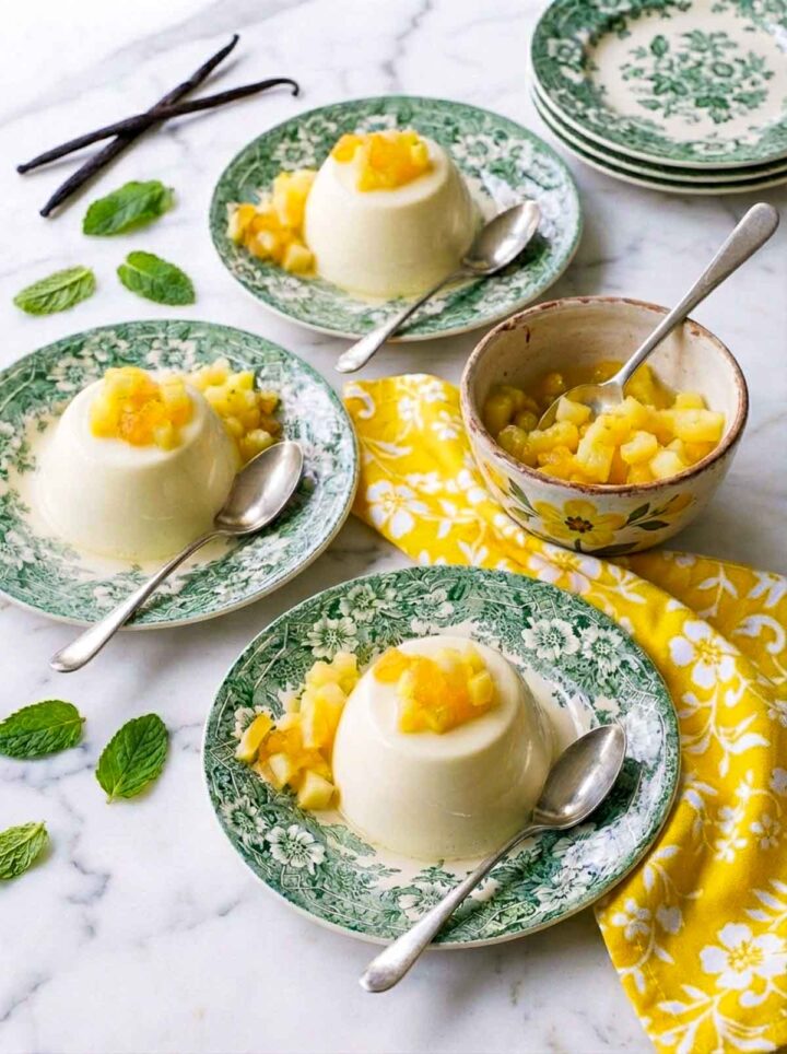 Vanilla bean panna cotta served on green patterned plates topped with fresh pineapple pieces, set on a marble surface with a bright yellow patterned napkin and mint leaves.