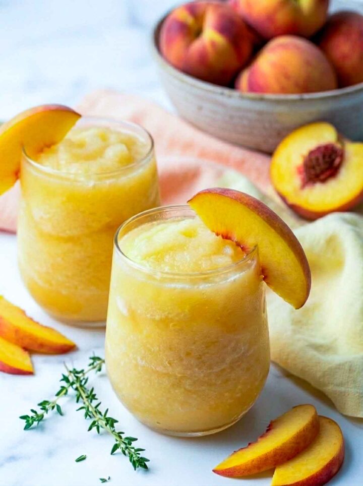  Two peach limoncello slush drinks in short rounded glasses garnished with peach slices, surrounded by fresh peaches, a bowl of whole peaches, and a soft peach-colored napkin on a marble surface.
