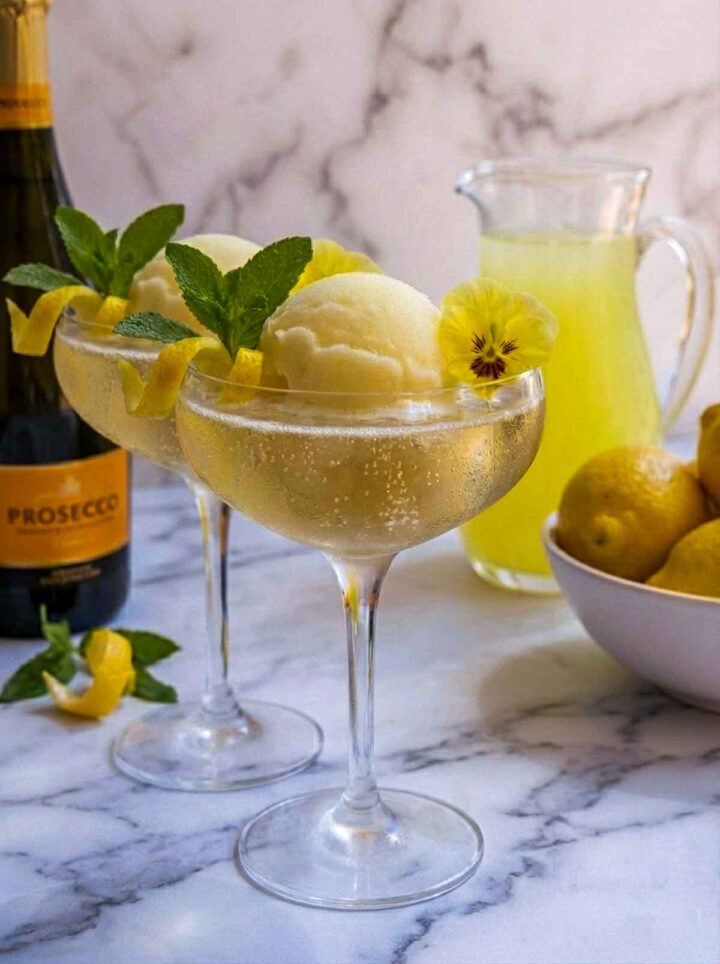 Two coupe glasses filled with limoncello and Prosecco with scoops of lemon sorbet, garnished with lemon peel twists, mint, and edible flowers, with a Prosecco bottle and lemons in the background.