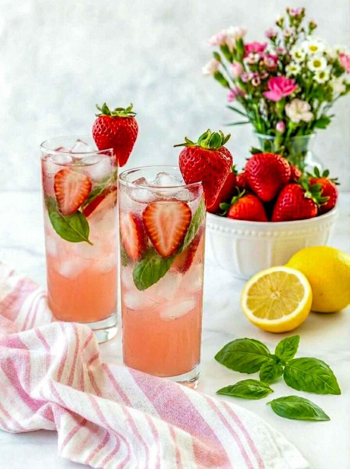  Two tall glasses of sparkling limoncello strawberry basil mocktail with sliced strawberries and basil leaves, garnished with whole strawberries on the rim, with lemons and fresh strawberries in the background on a marble countertop.