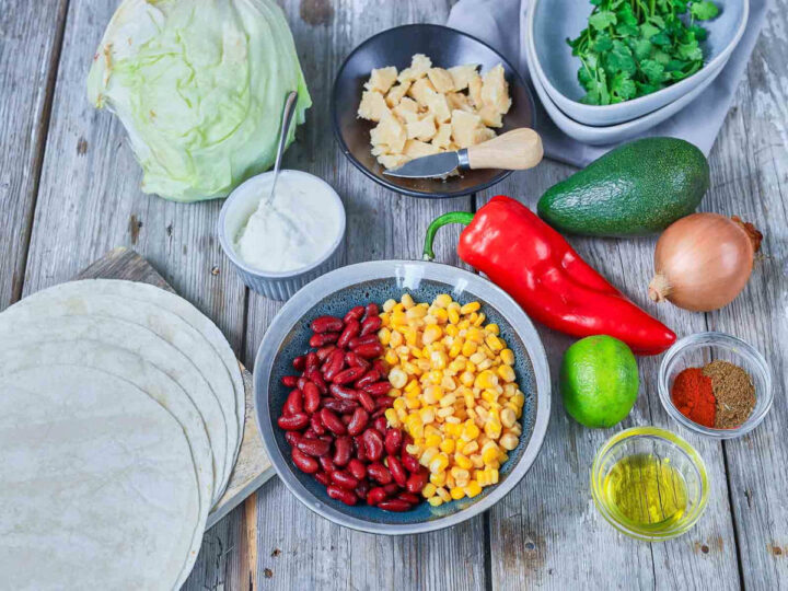 Fresh ingredients like beans, corn, cabbage, avocado, pepper, onion, lime, herbs, spices, and tortillas are prepared for a vibrant salad.