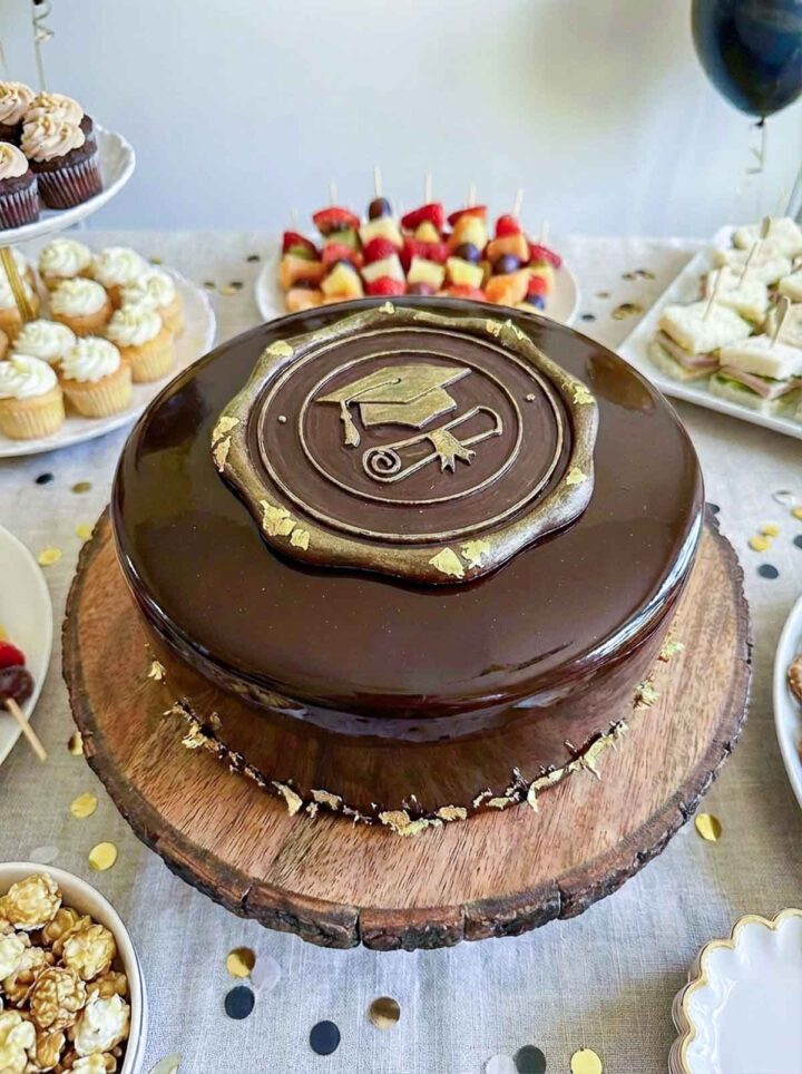 Chocolate mirror glaze cake placed on a cake stand with other party food surrounding it.