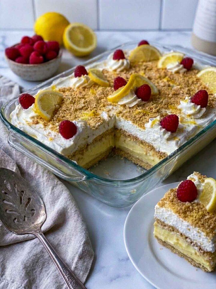  No bake limoncello lemon icebox cake in a glass dish with layered lemon cream and wafers, topped with whipped cream, crushed crumbs, lemon slices, and raspberries, with a slice served on a plate.