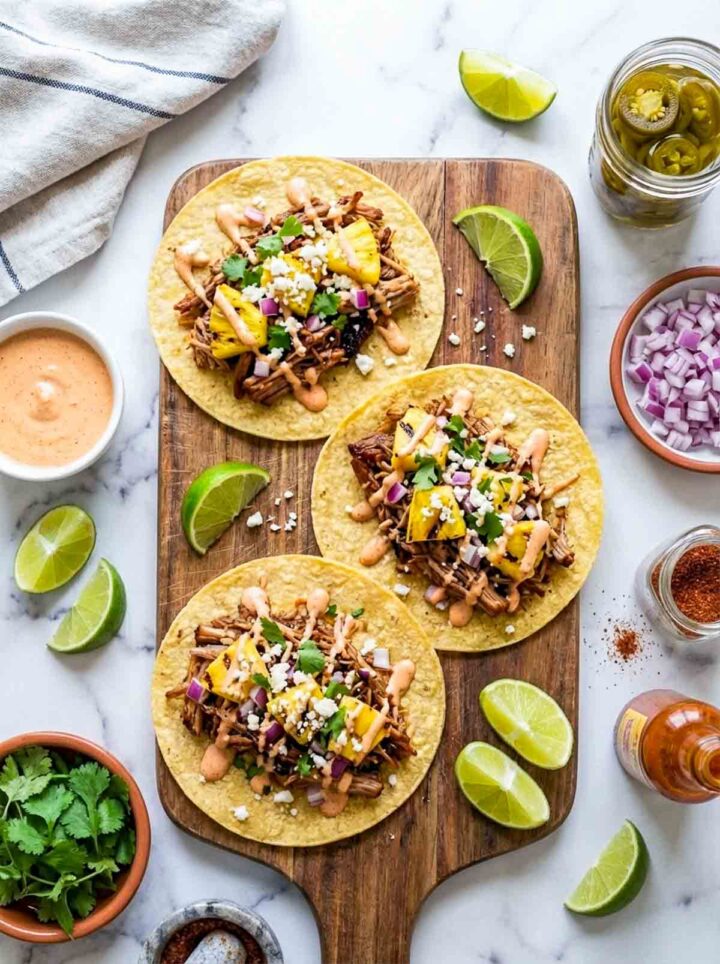 Pineapple chipotle pulled pork tacos topped with diced pineapple, red onion, cilantro, and sauce, served with lime wedges on a wooden board over a marble surface.