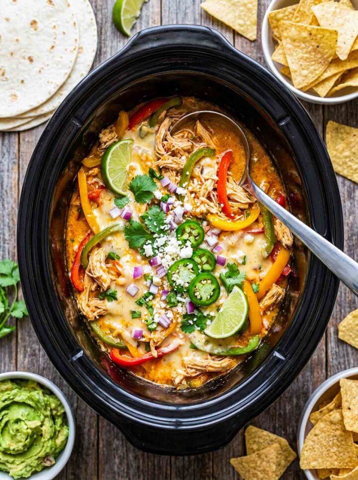 Slow cooker queso chicken with shredded chicken, melted cheese, and peppers, topped with jalapenos, onion, and cilantro, served with tortillas and chips.