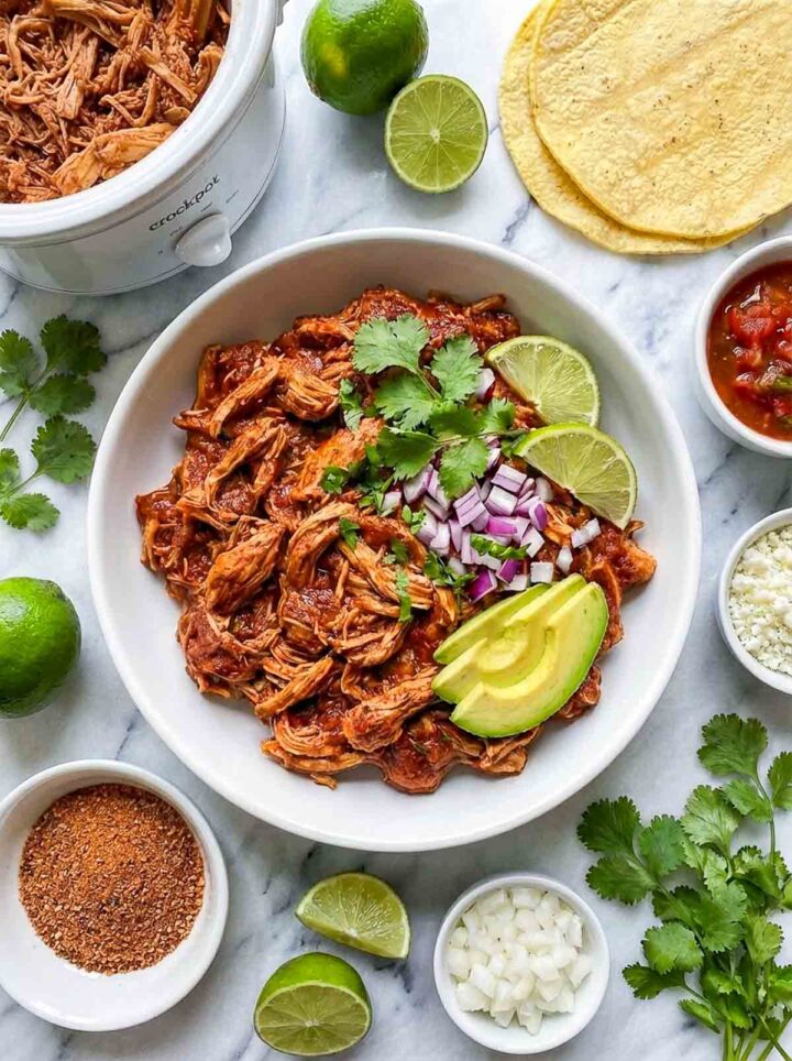 Tajin lime shredded chicken in a bowl topped with avocado, onion, cilantro, and lime, served with tortillas and seasoning on a marble surface.