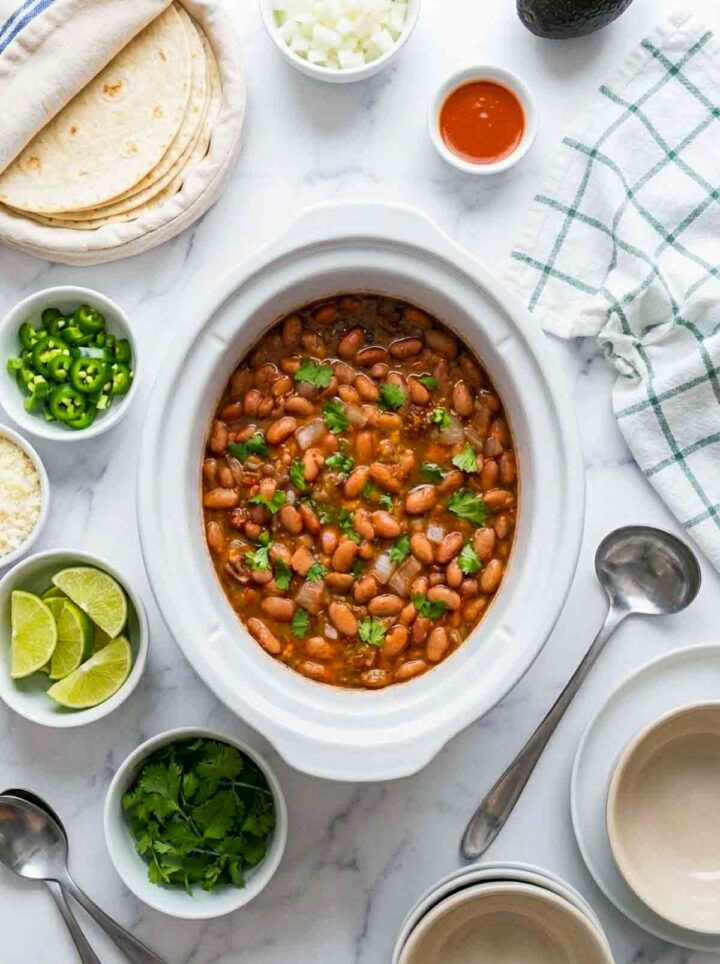 Slow cooker Mexican pinto beans in a crockpot with cilantro, served with tortillas, lime, jalapenos, and taco bar toppings on a marble surface.