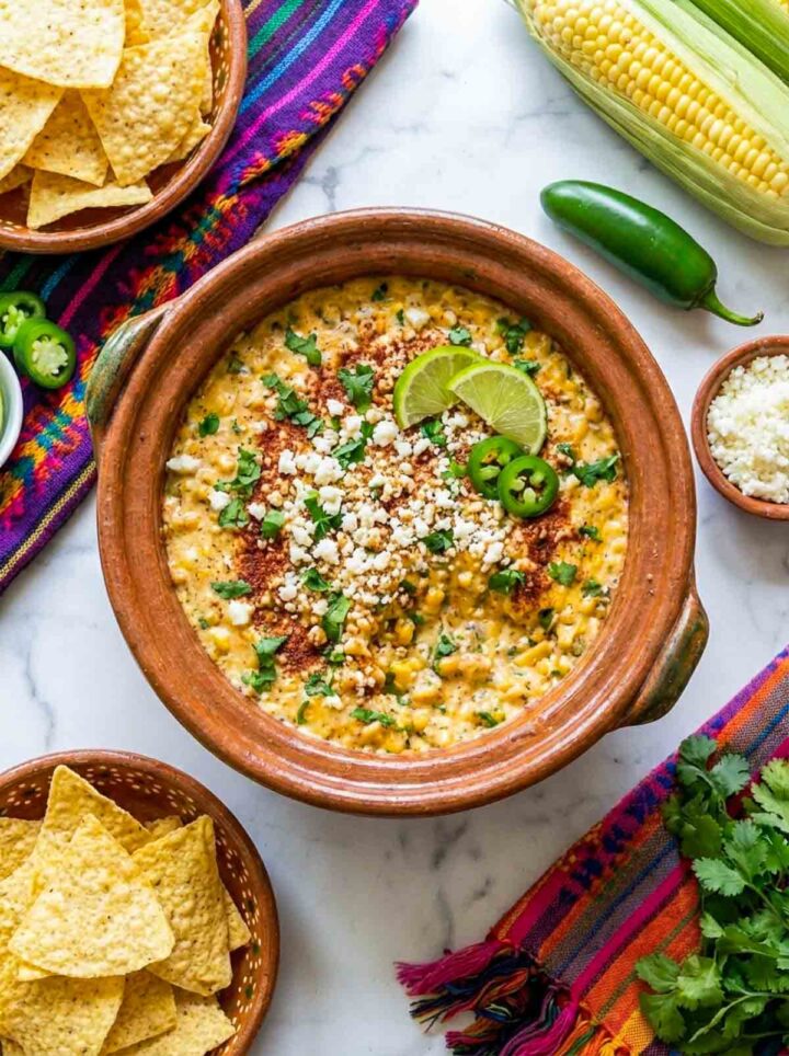 Mexican street corn dip with chicken topped with cotija cheese, chili powder, cilantro, and lime, served with tortilla chips on a marble surface.