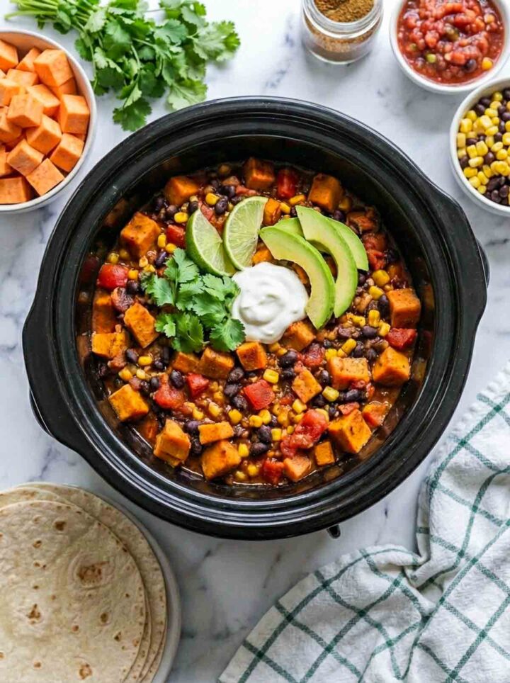 Sweet potato black bean taco filling in a crockpot topped with avocado, cilantro, and sour cream, served with tortillas on a marble surface.