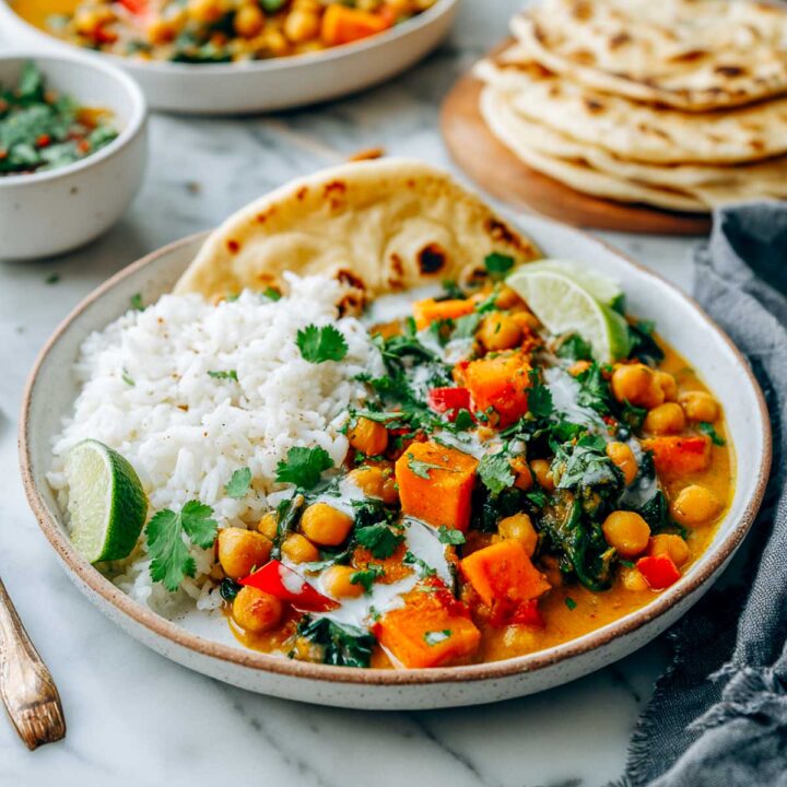 A plate of sweet potato chickpea curry served with white rice, naan, lime wedge, and fresh cilantro on top.