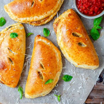 Four golden-brown chicken pesto calzones arranged on parchment paper with fresh basil leaves, grated Parmesan, and a bowl of marinara dipping sauce.