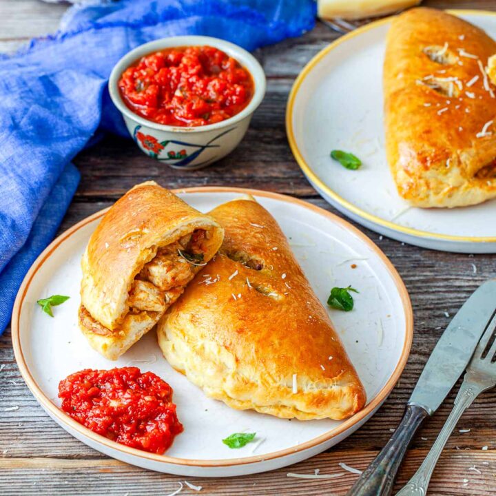 Chicken pesto calzones served on plates, including one cut open to reveal the chicken filling, with marinara sauce for dipping.