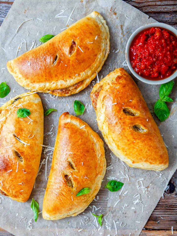 Freshly baked chicken pesto calzones with crisp golden crusts, garnished with basil leaves and Parmesan, served alongside marinara sauce.