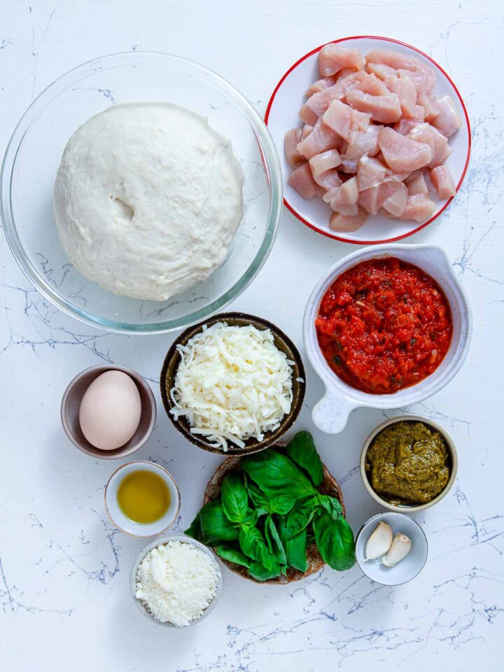 Ingredients for chicken pesto calzones including pizza dough, chicken breast, marinara sauce, pesto, mozzarella, Parmesan, basil, garlic, egg, and olive oil.