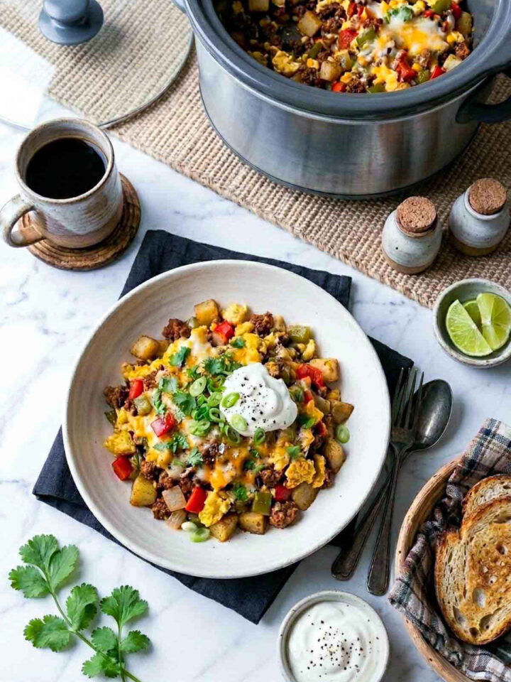 Overhead shot of a crockpot Mexican breakfast casserole served in a wide speckled ceramic bowl, showing diced potatoes, crumbled chorizo, scrambled eggs, red and green peppers, and melted cheese, topped with a dollop of sour cream, sliced green onions, and fresh cilantro, with an unbranded slow cooker in the background.