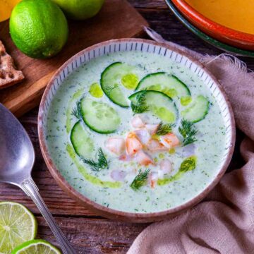 Overhead-angle view of chilled cucumber avocado soup in a ceramic bowl, topped with cucumber slices, dill sprigs, and optional cumin-seasoned shrimp garnish on a rustic wood surface.