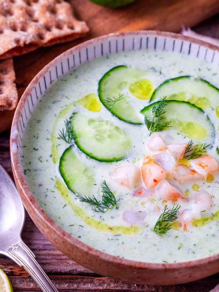 Close-up of chilled cucumber avocado soup in a ceramic bowl topped with overlapping cucumber slices, fresh dill sprigs, and optional cumin-seasoned shrimp garnish.